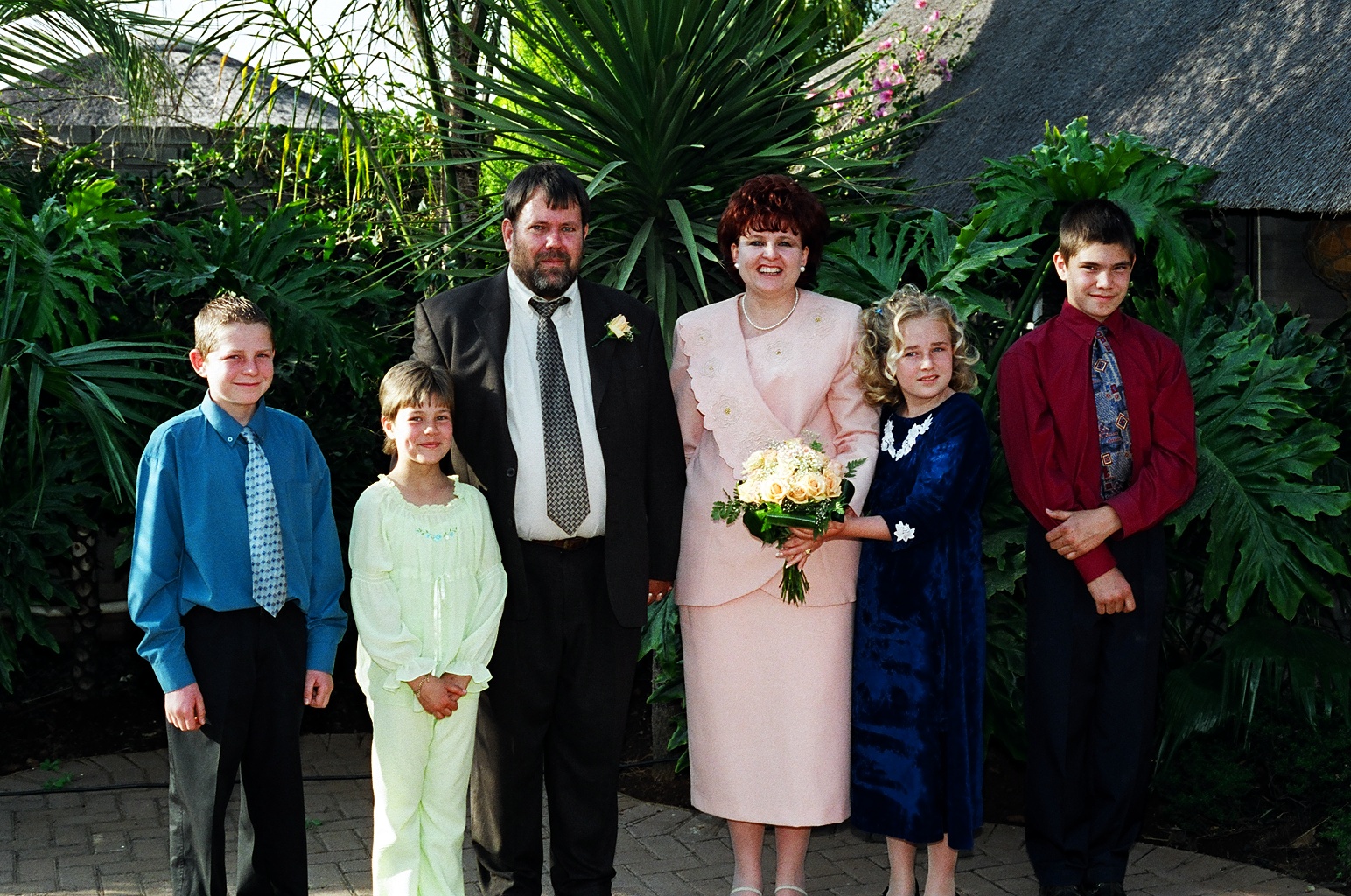 Family photograph from a wedding day: young Angelo in a blue shirt and tie standing with his family in front of palm trees