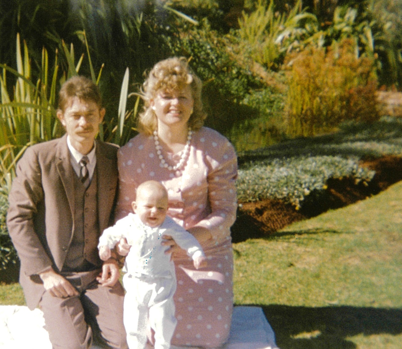 A couple kneeling on a white blanket on the lawn with a baby in a white christening outfit