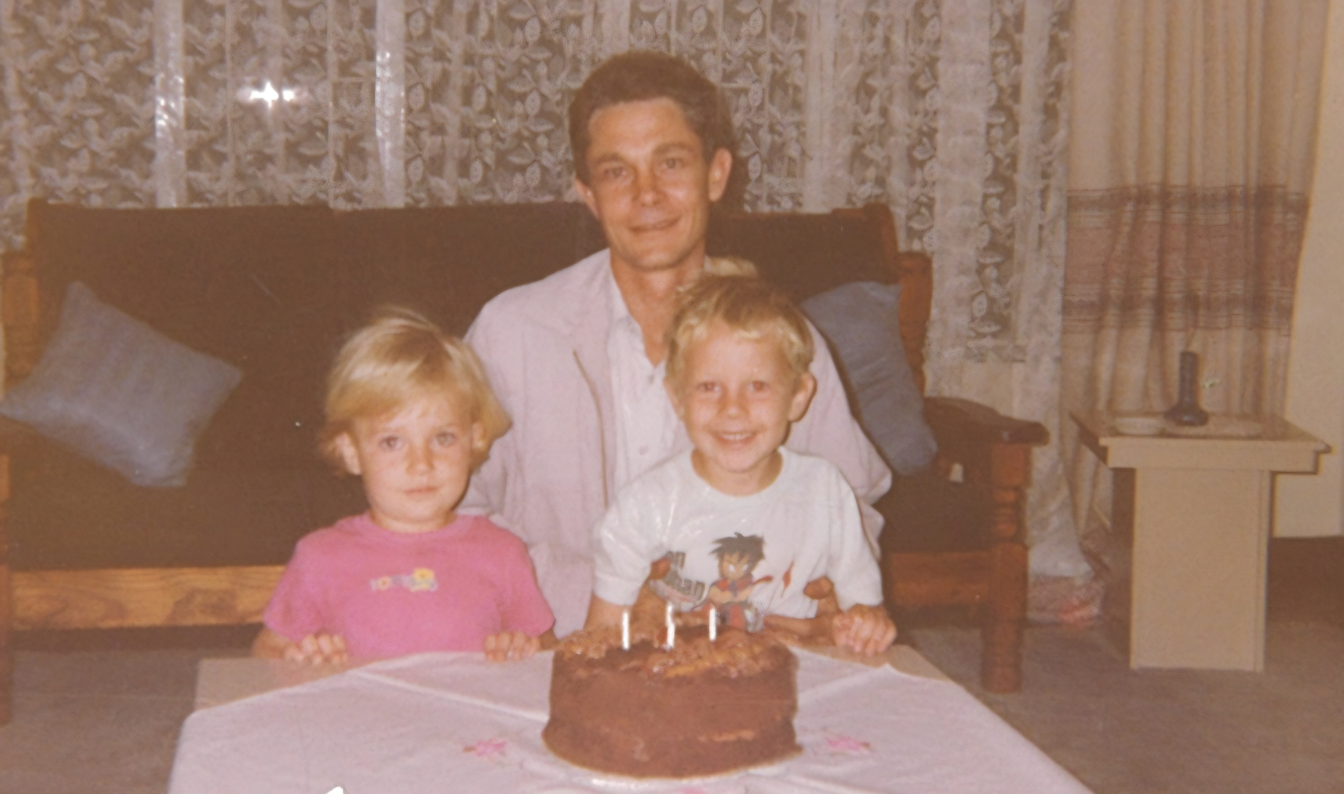A man and two children seated behind a chocolate birthday cake with lit candles