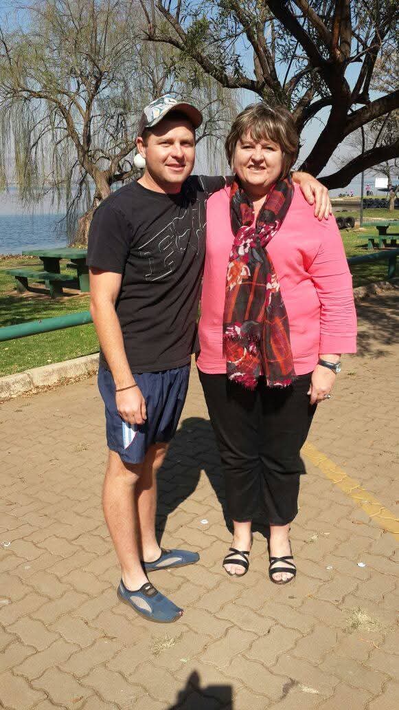 Angelo with his arm around his mum on a paved path by the water