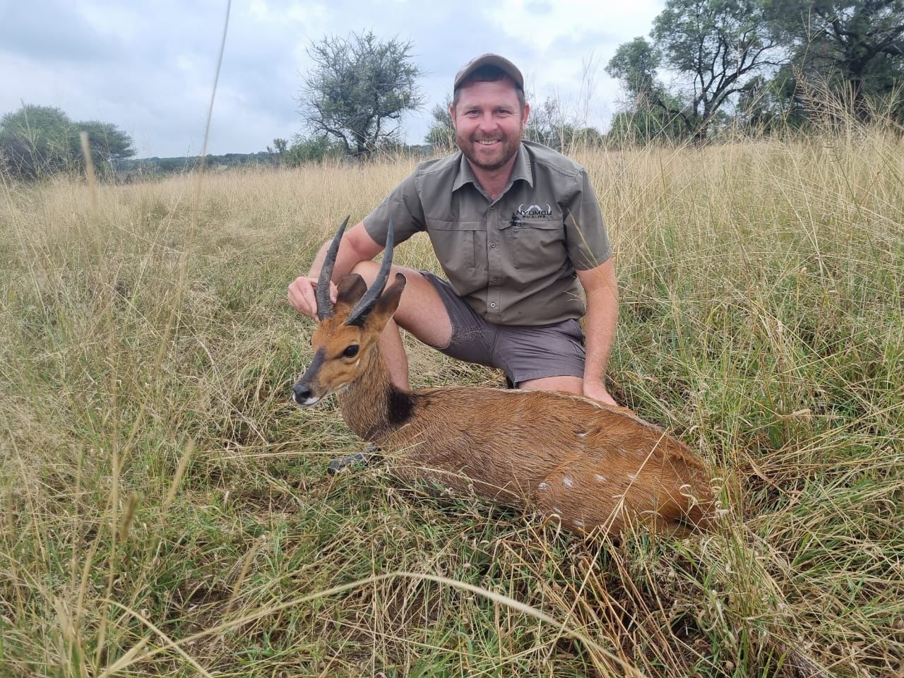 Angelo kneeling in tall grass beside a bushbuck ram