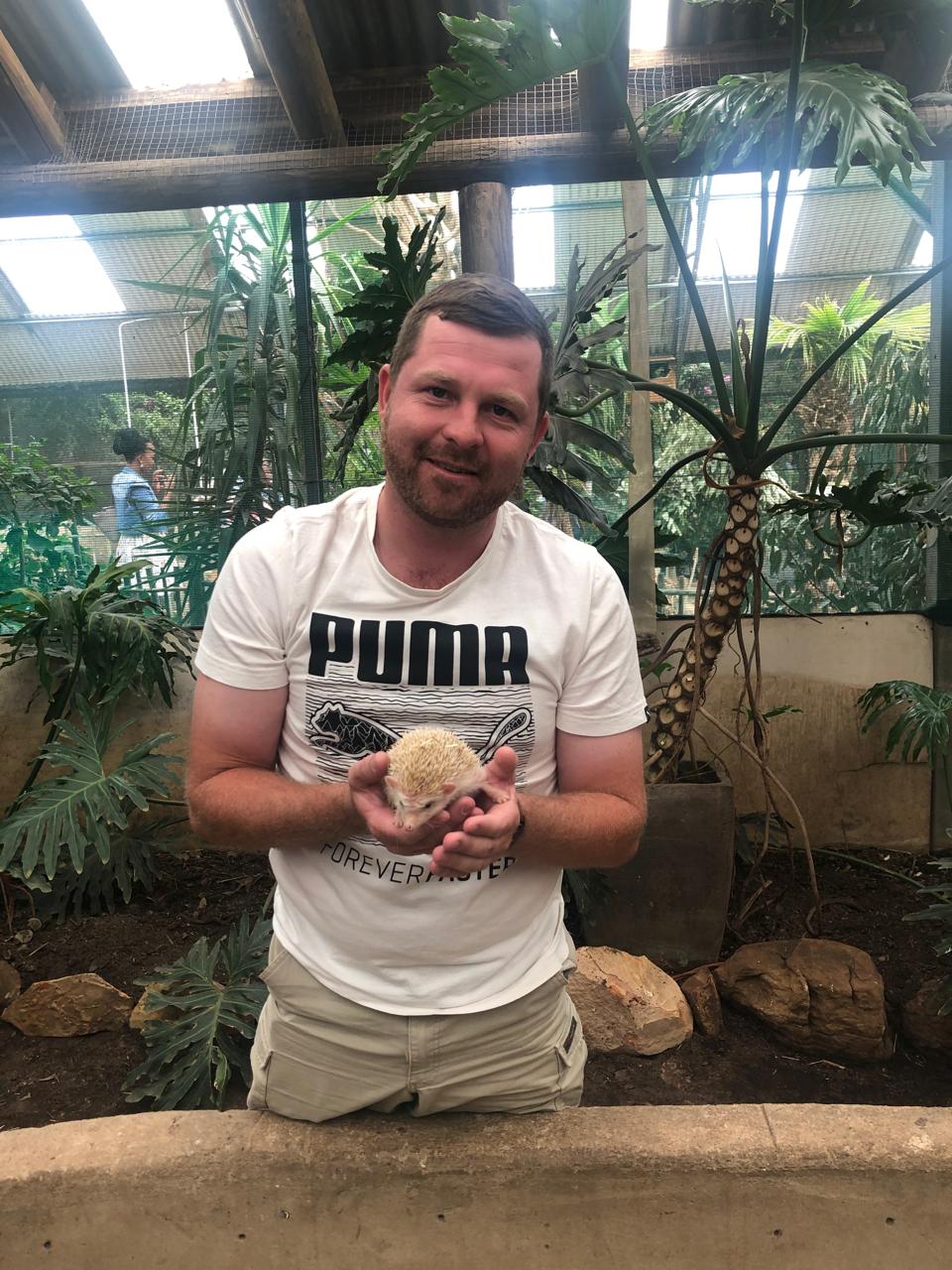 Angelo kneeling in a greenhouse, smiling while holding a small hedgehog in his hands