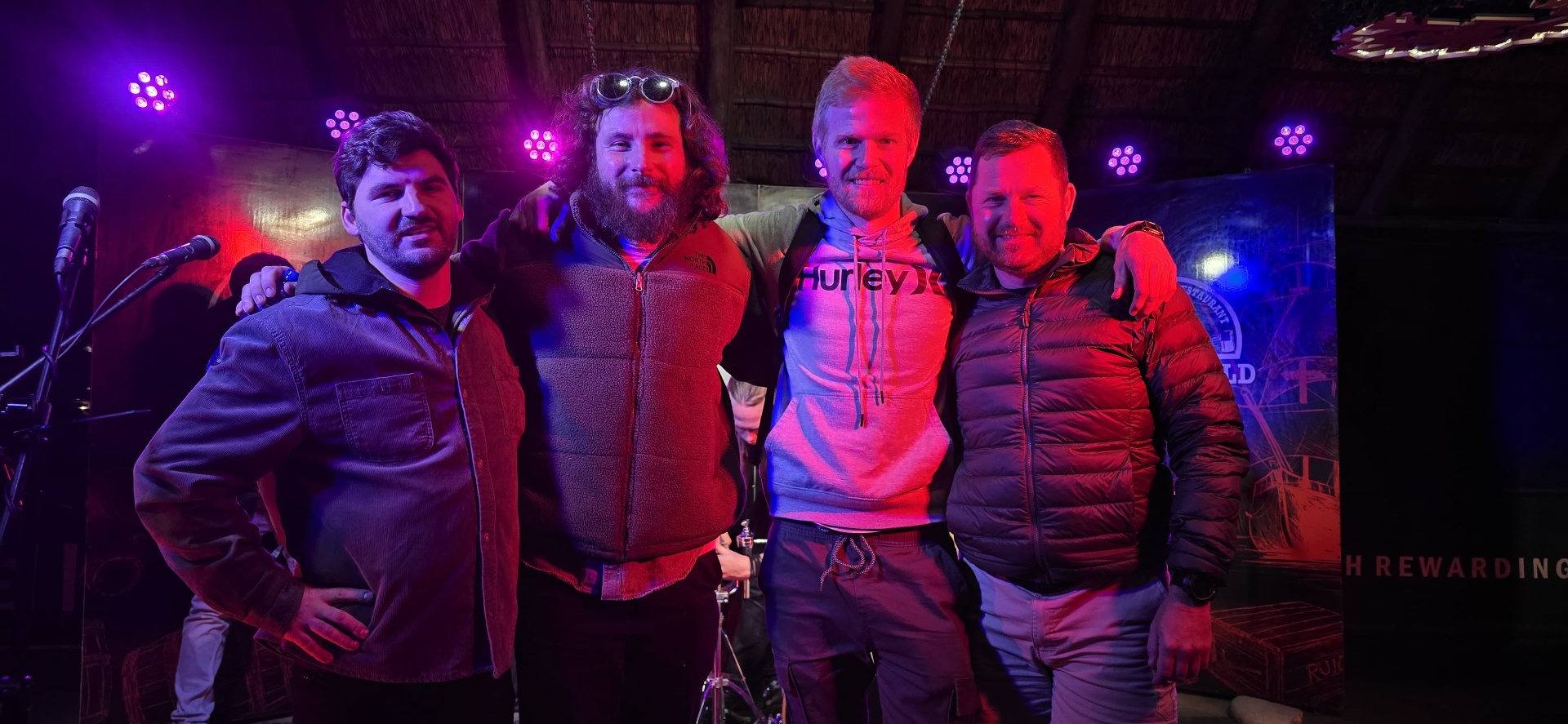 Four friends standing arm in arm on a stage beneath coloured lights