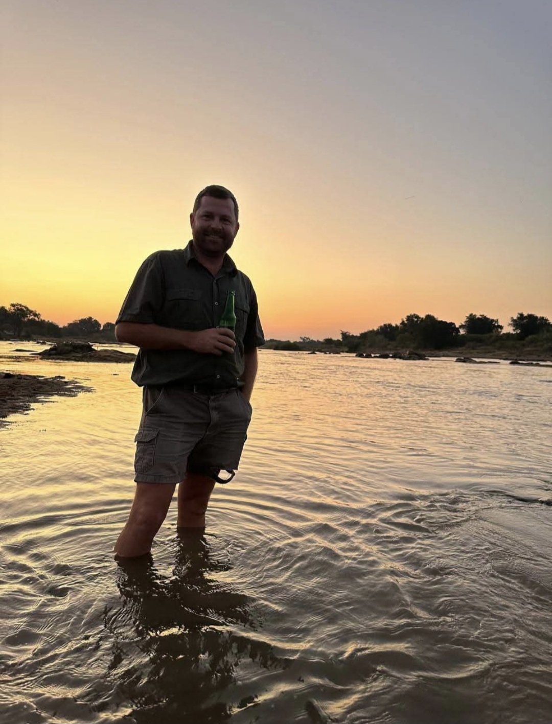 Angelo standing knee-deep in a river at sunset, holding a bottle and smiling