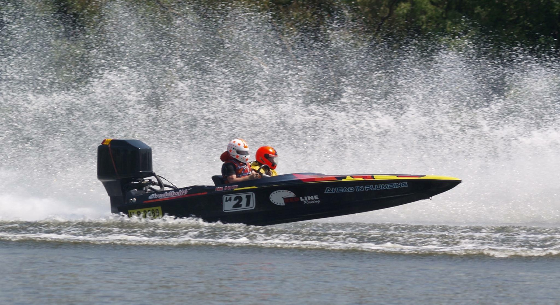 Two helmeted racers in a black number 21 boat speeding past a wall of spray