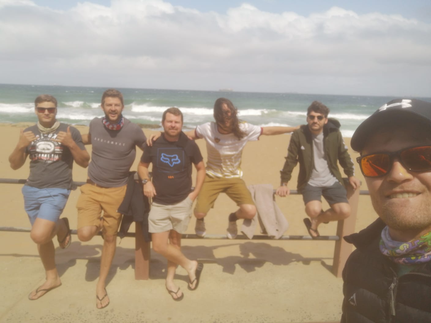 Angelo and five friends leaning on a railing above a golden beach