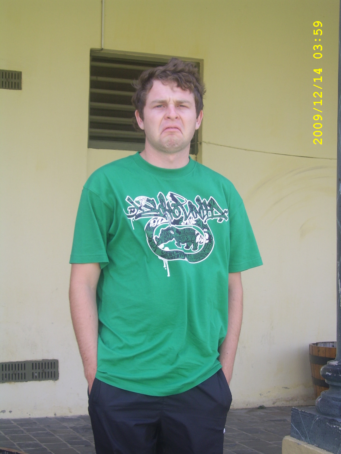 A young Angelo in a green shirt making a playful expression beside a yellow building