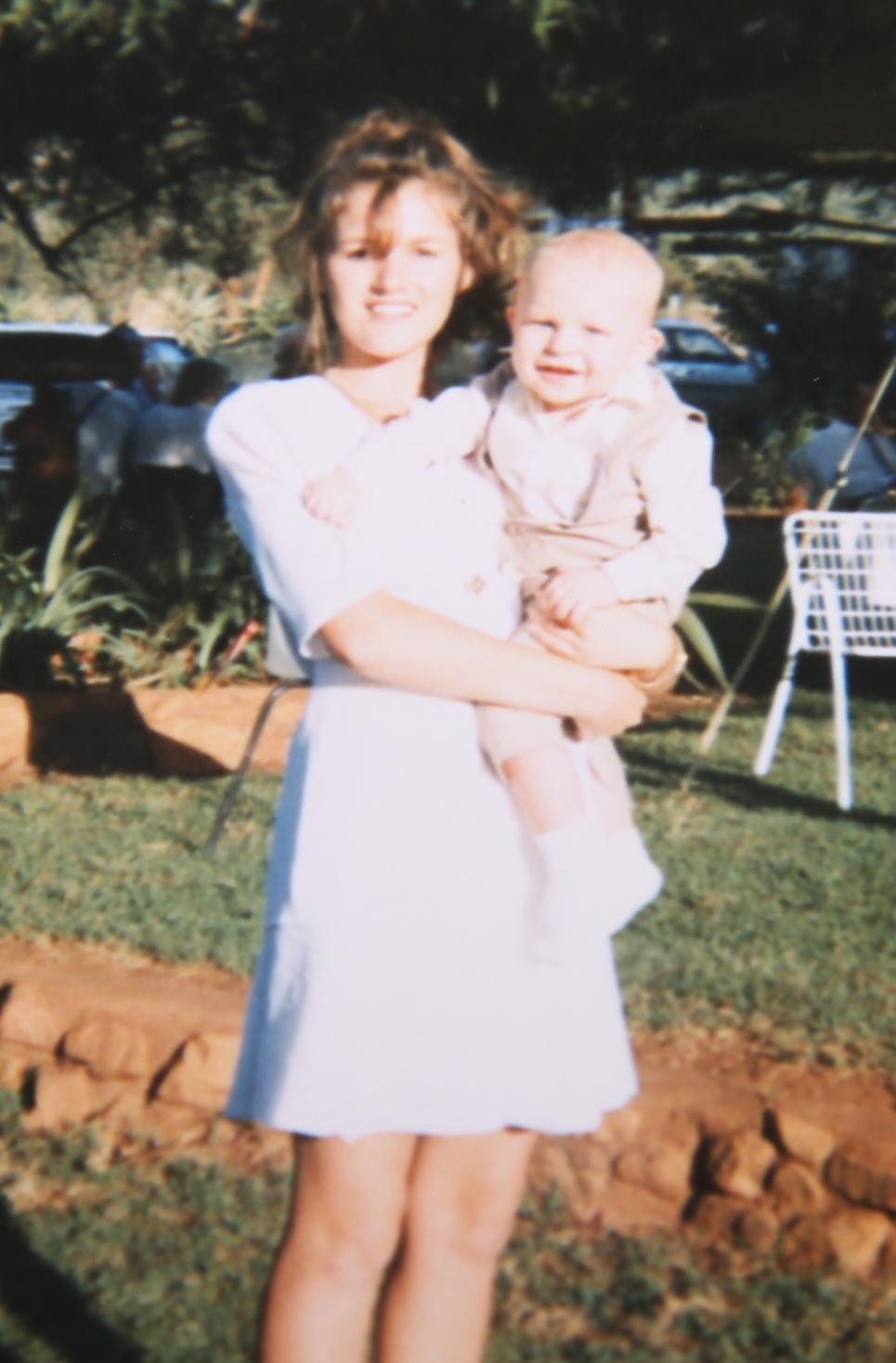 A young woman holding a smiling baby in a little waistcoat on a sunny lawn