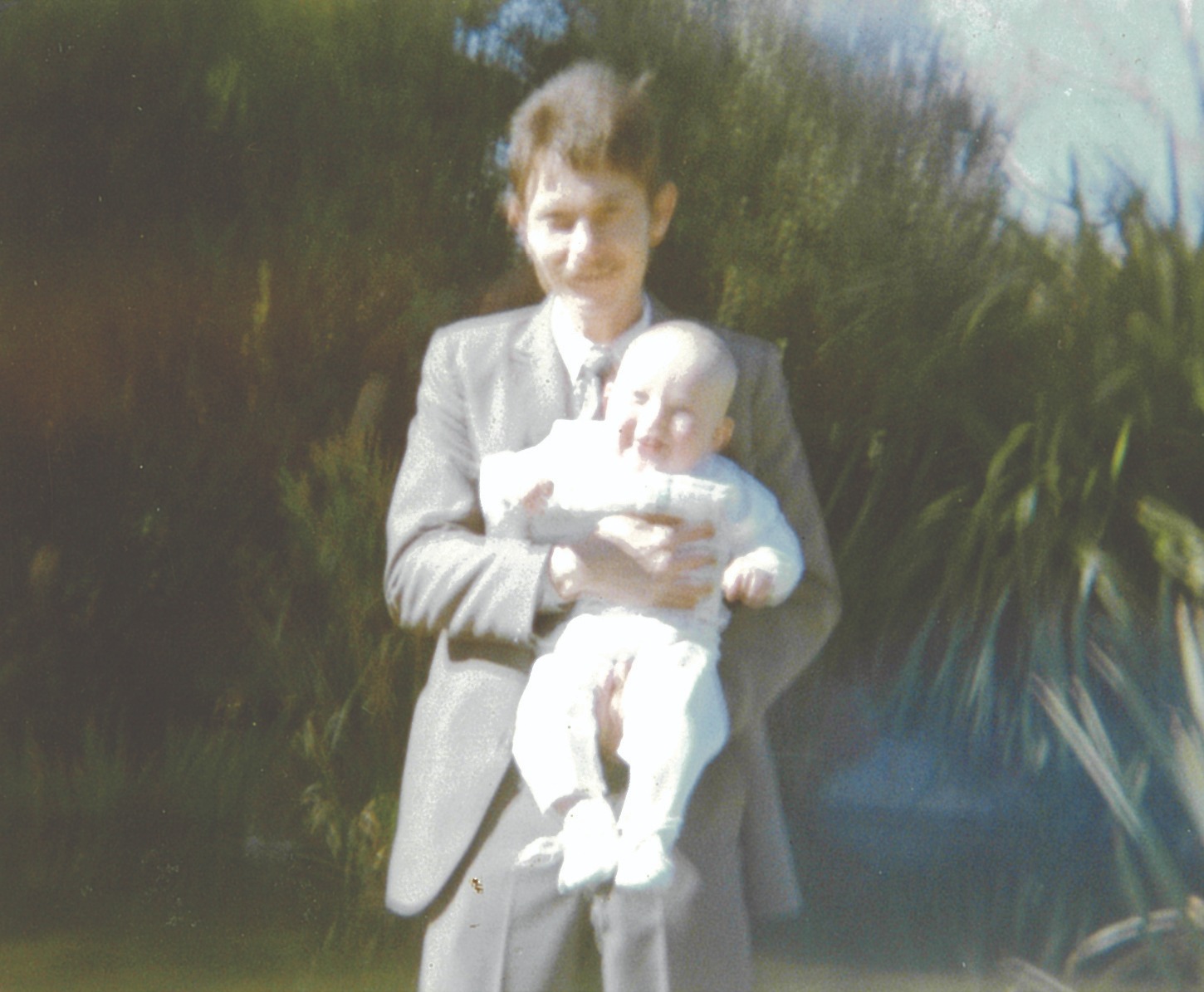 A man in a suit holding a baby in a white christening outfit among garden bushes