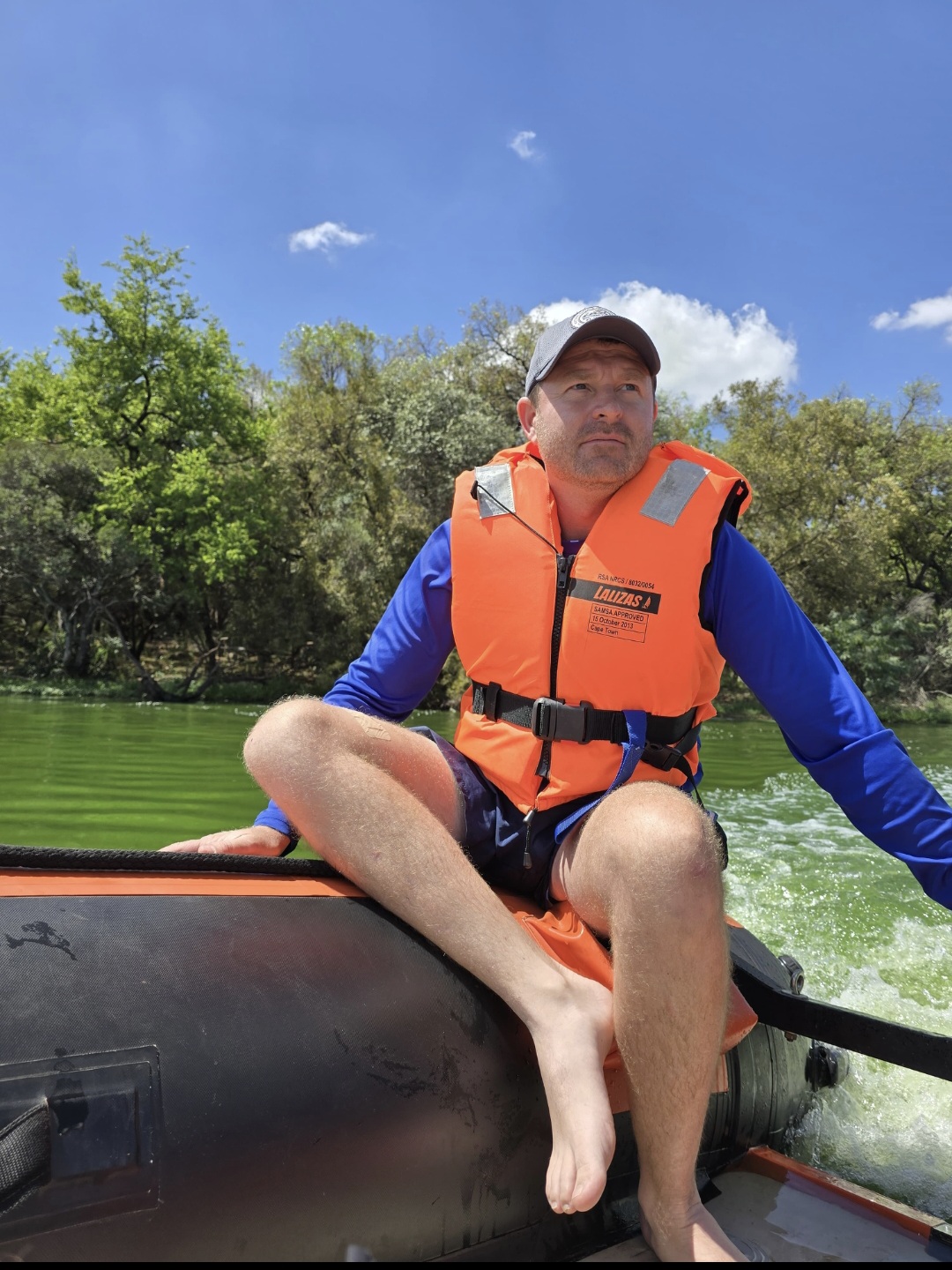 Angelo sitting on the edge of an inflatable boat on a green river wearing an orange life jacket