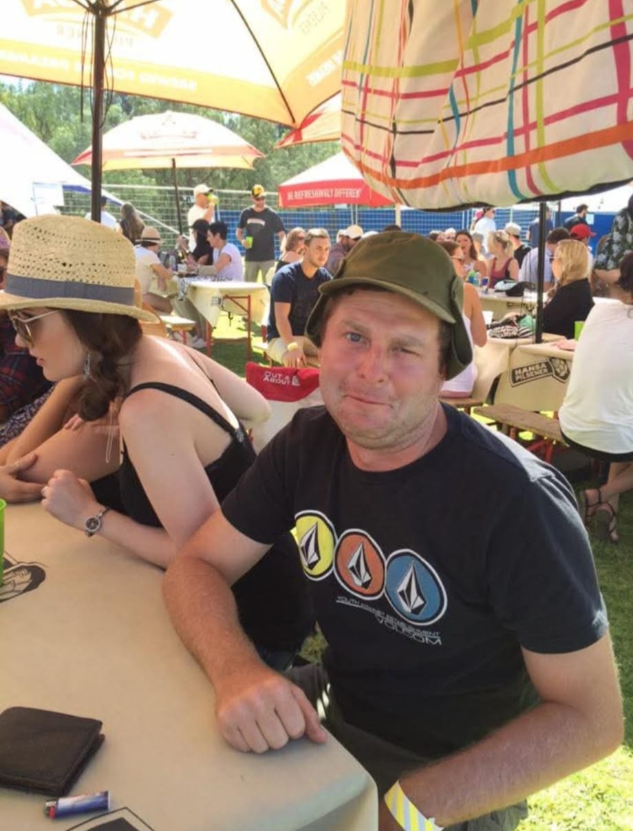 Angelo pulling a playful face at a festival table under a striped umbrella