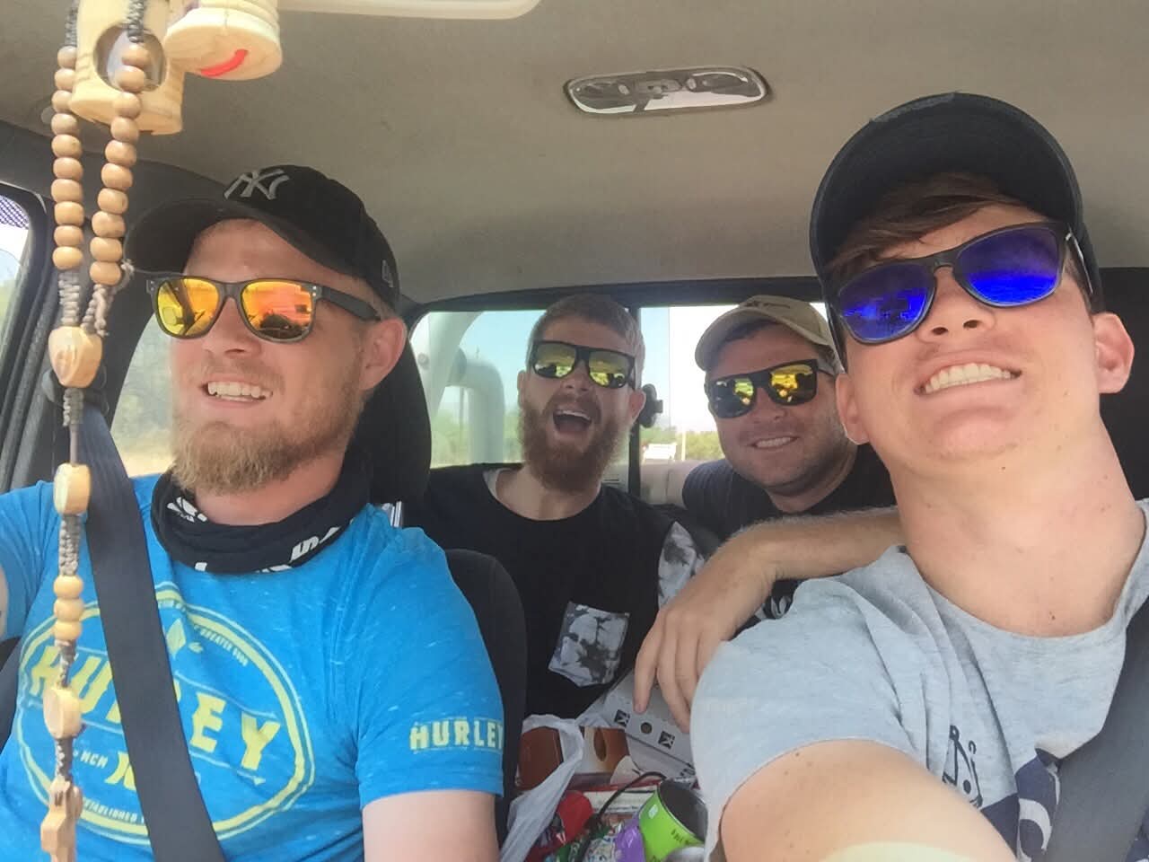 Four friends in sunglasses smiling in a vehicle selfie on a road trip