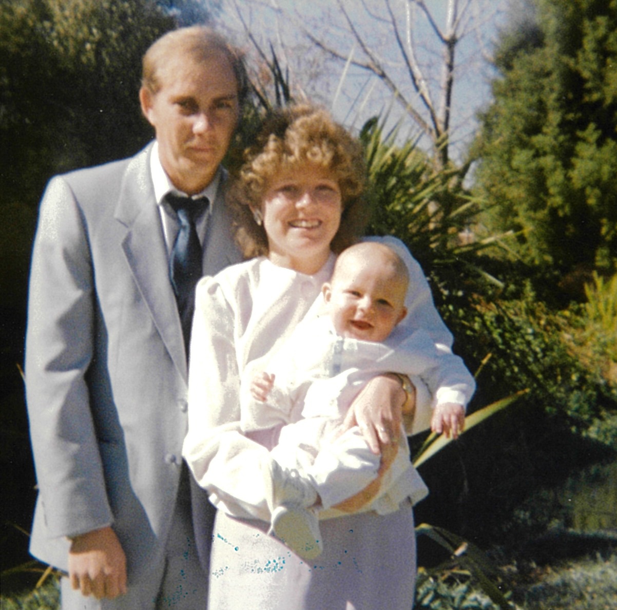 A family in formal clothes posing outdoors with a baby in a white christening outfit