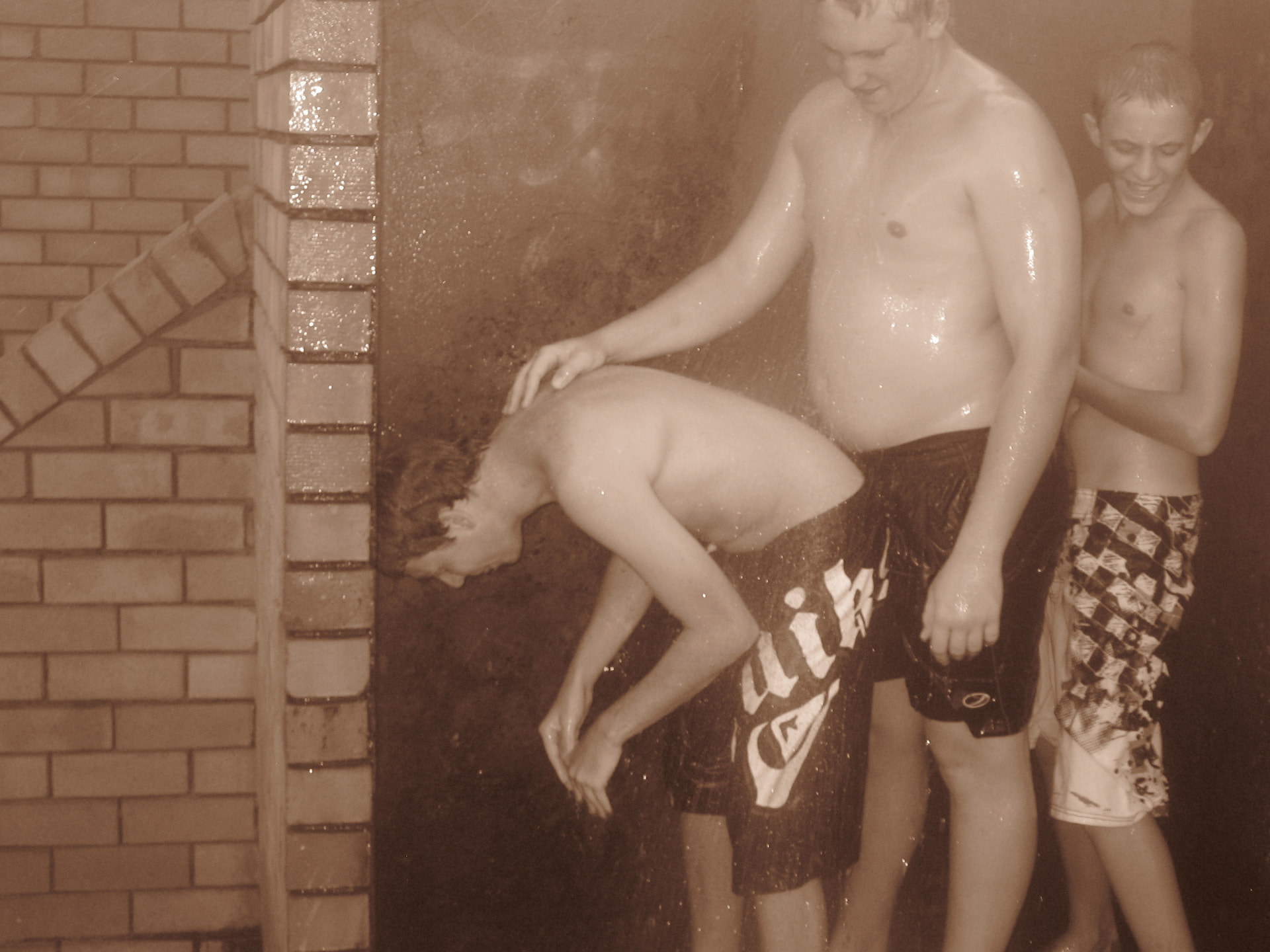 A sepia photograph of Angelo and friends sharing a playful moment beside a brick outdoor shower at church camp