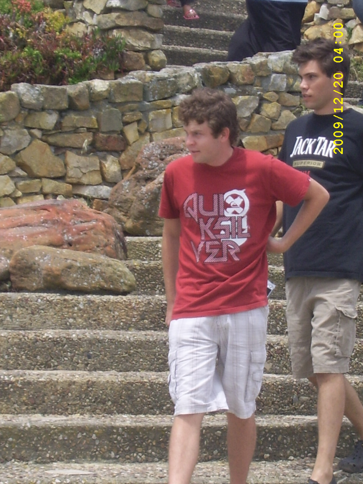 A young Angelo in a red T-shirt on stone steps beside a friend and a rock wall