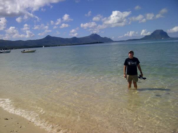 Angelo standing knee-deep in clear turquoise shallows on a sandy beach