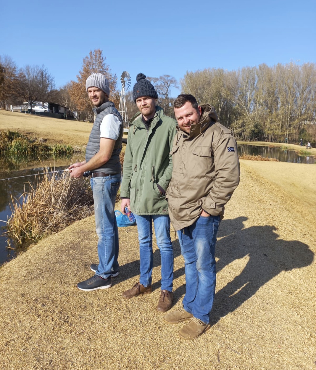 Three friends in winter jackets beside a dam with a fishing rod