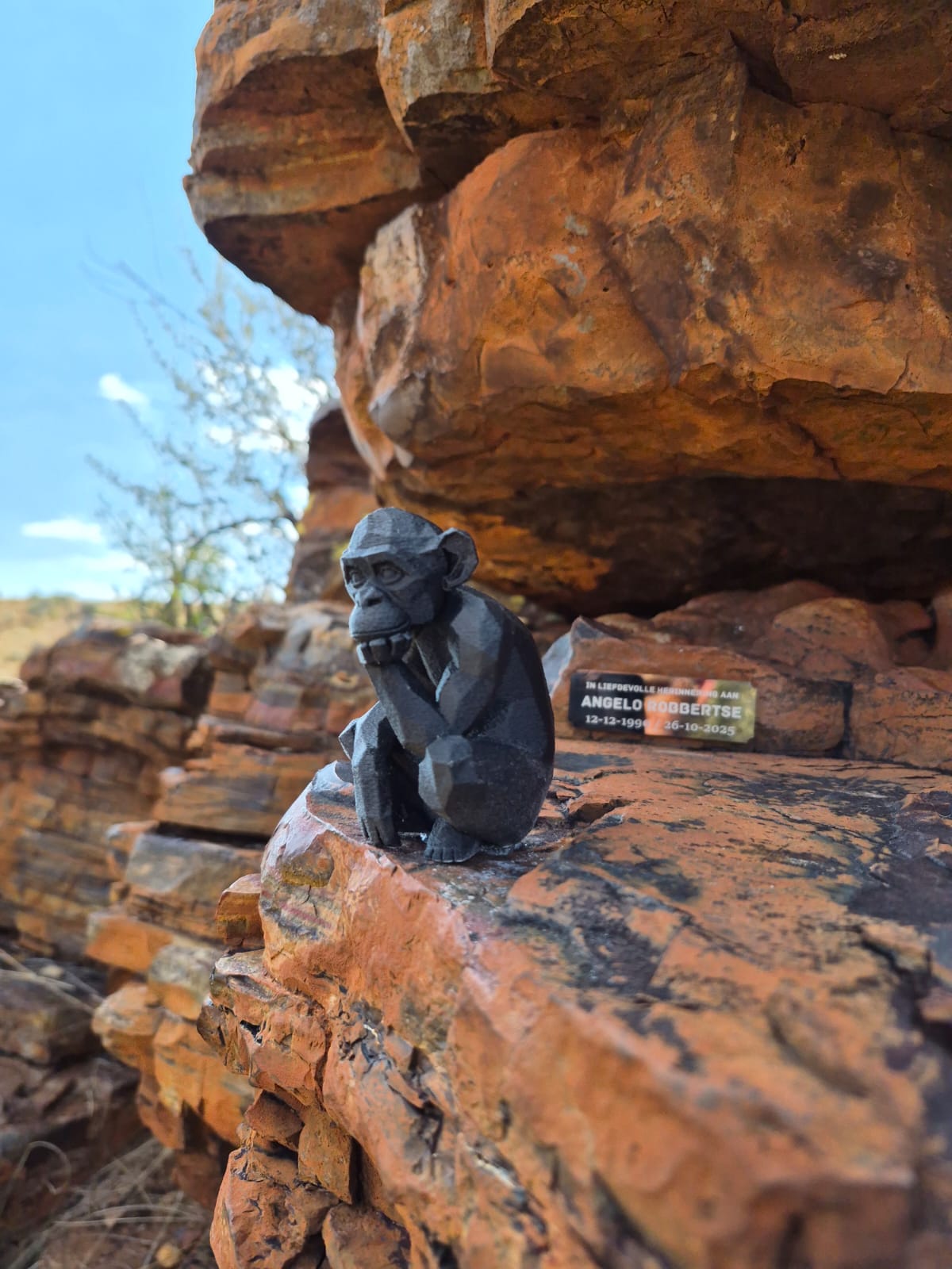 A small carved monkey figure resting on red rock beside Angelo's memorial plaque