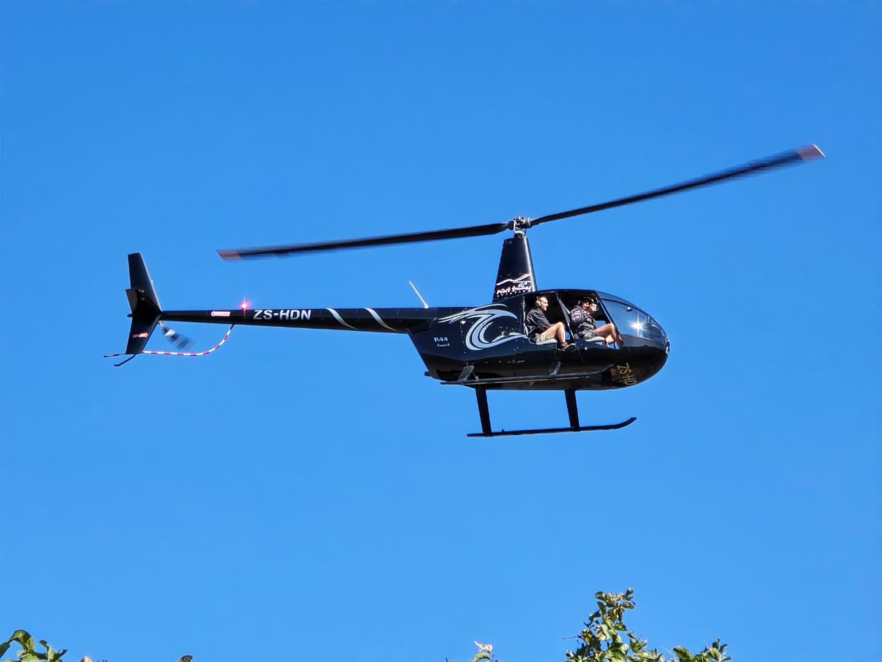 A black ZS-HDN helicopter flying beneath a clear blue sky with two people aboard