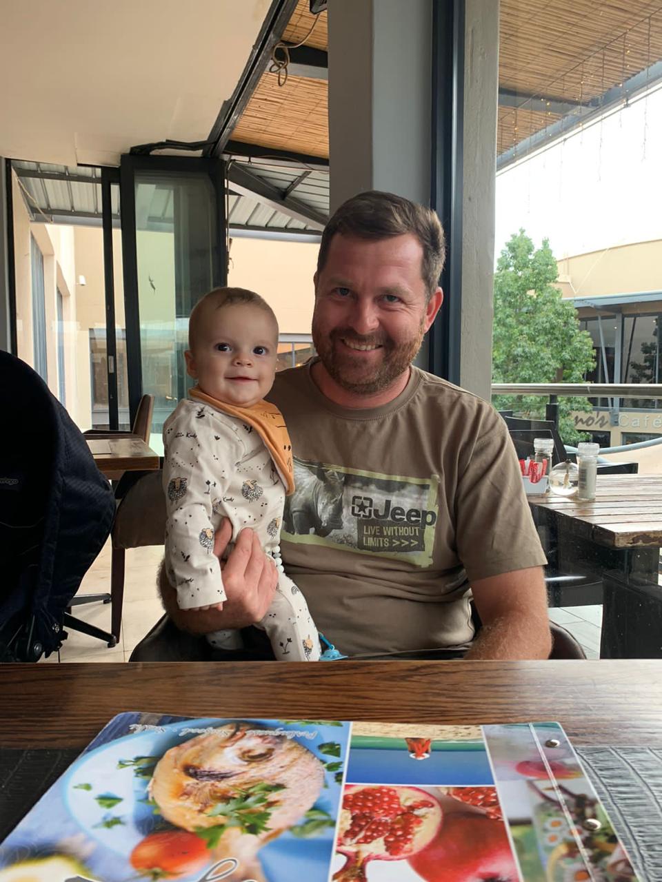 Angelo smiling with a baby on his lap at a cafe table