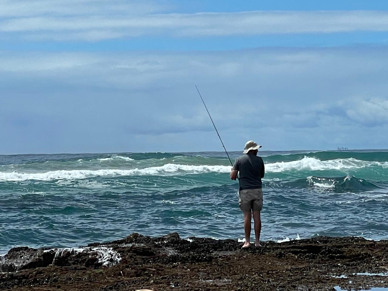 Angelo fishing with a rod from the rocks as green ocean waves roll in