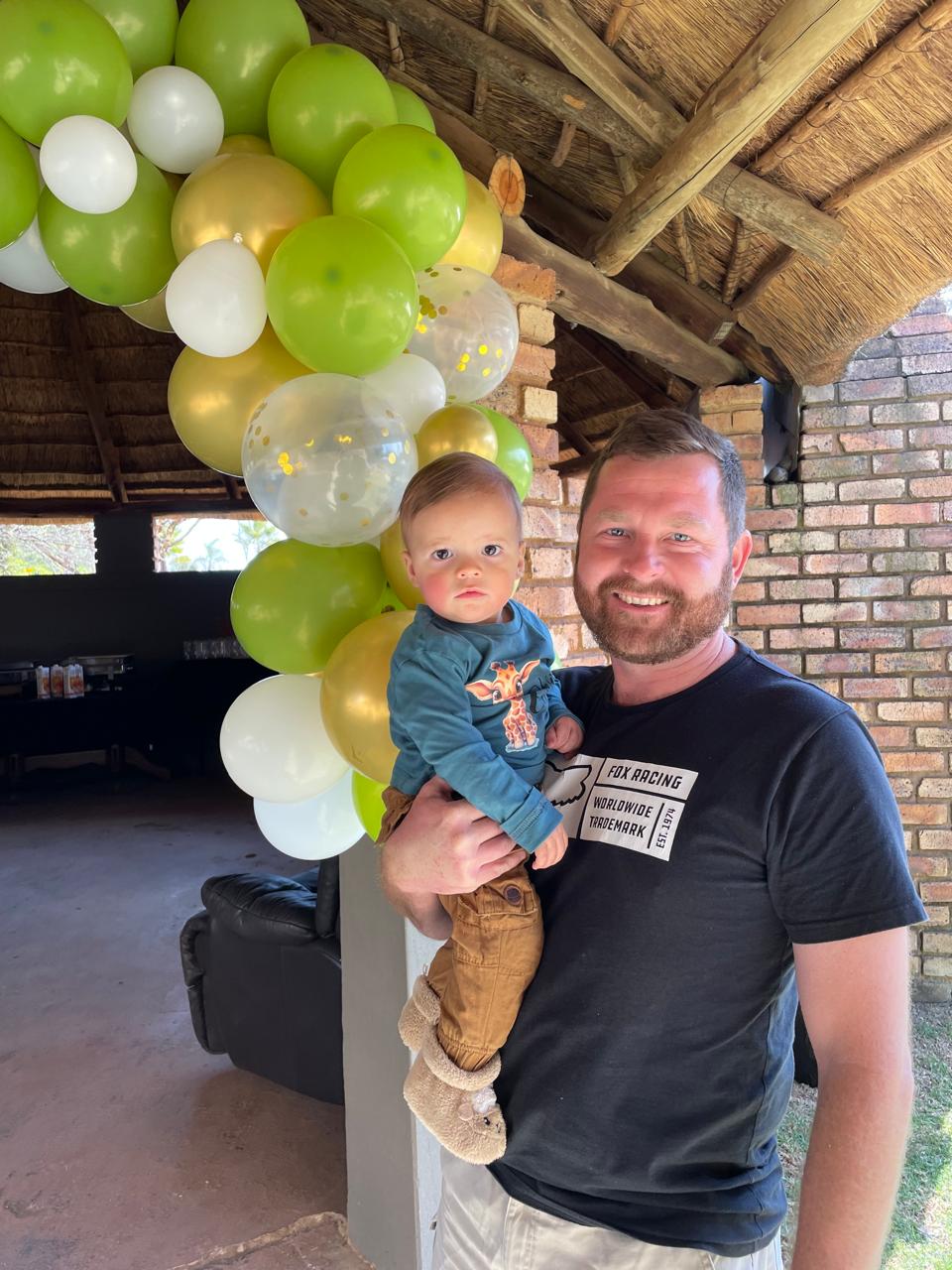 Angelo holding a baby in front of a green and gold balloon arch