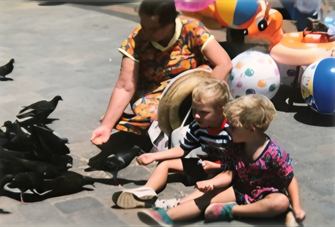 A woman and two young boys sitting on a stone pavement feeding a flock of pigeons
